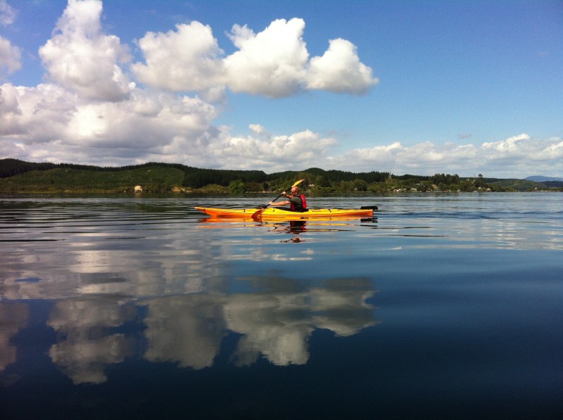 Person kayaking on calm water with cloudy sky reflection.
