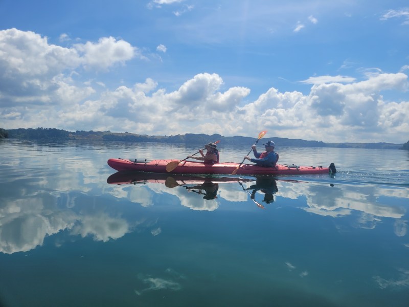 Two people kayaking on a calm lake under a blue sky with clouds.